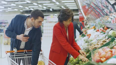 En-el-supermercado:-feliz-pareja-realiza-compras,-elegir-frutas-y-verduras-en-fresco-producen-sección.-Hombre-utiliza-Smartphone-y-empuja-el-carrito-de-compras,-mujer-coloca-productos-en-carro.