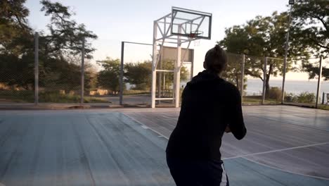 Vista-rara-de-un-tiro-de-larga-distancia-por-el-jugador-de-baloncesto-femenino-joven-practicando-en-la-cancha-de-baloncesto-al-aire-libre-local.-Árboles-y-el-sol-brilla-en-el-fondo