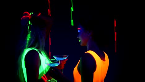 Women-with-UV-face-paint,-laser,-glowing-bracelets,-drinks,-glowing-clothing-dancing-together-in-front-of-camera,-Half-body-shot.-Caucasian-and-asian-woman.-.