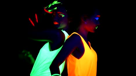 Women-with-UV-face-paint,-glowing-bracelets,-glowing-glasses,-clothing-dancing-back-against-each-other-in-front-of-camera,-Half-body-shot.-Caucasian-and-asian-woman.-Women.