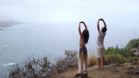 Un-hombre-y-una-mujer-de-pie-en-el-borde-de-un-acantilado-con-vistas-al-océano-levantan-sus-manos-y-aspirar-el-aire-del-mar-durante-yoga