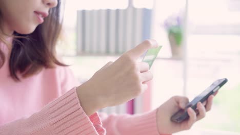 Beautiful-Asian-woman-using-smartphone-buying-online-shopping-by-credit-card-while-wear-sweater-sitting-on-desk-in-living-room-at-home.-Lifestyle-woman-at-home-concept.