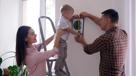 home-repair,-cute-child-with-measuring-tape-helps-to-parents-make-repairs-and-hang-picture-in-apartment