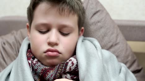 A-cold-boy-coughs.-He-has-the-flu,-he's-sitting-on-the-couch-in-a-scarf