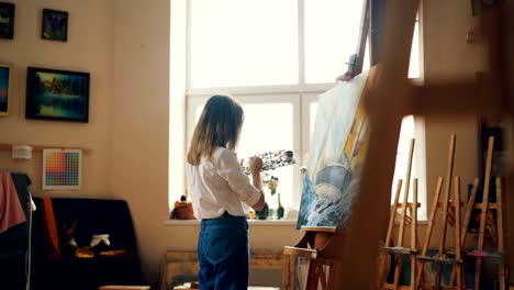 Professional-painter-young-lady-is-painting-seascape-with-acrylic-paints-depicting-marine-landscape-ship-and-sea-waves-working-alone-in-studio.-People-and-work-concept.