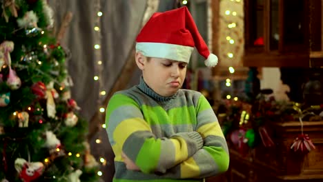 The-boy-in-the-sweater-and-the-hat-of-Santa-Claus-is-upset-and-offended.-He-has-no-gift.-against-the-background-of-Christmas-lights