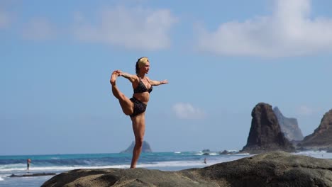 Young-girl-in-bikini-balancing-stands-on-one-leg-doing-yoga-standing-on-a-rock-on-the-ocean-beach-with-black-sand.-Meditation-through-relaxation.-Gymnastics