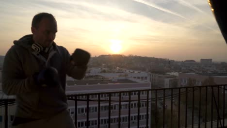 Mann-mit-Handschuhen-an-den-Händen-Schläge-Tasche-auf-Boxtraining-auf-der-Terrasse-im-Sonnenuntergang,-Sportler-üben,-Kraft-Training,-starken-Kerl-hart-trainieren,-Kraft-Übungen,-Training,-handheld,-sonnigen-Tag.