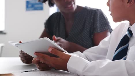 Close-Up-Of-Female-High-School-Tutor-With-Male-Student-Wearing-Uniform-Using-Digital-Tablets-For-One-To-One-Tuition-In-Classroom