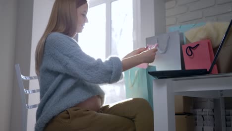 care-of-future-child,-happy-maternity-girl-with-bare-belly-examines-clothes-Internet-buying-for-an-future-baby-sitting-in-front-of-laptop