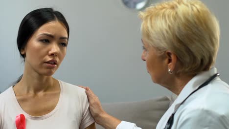 Sad-woman-with-pink-ribbon-visiting-doctor,-discussing-symptoms-of-breast-cancer