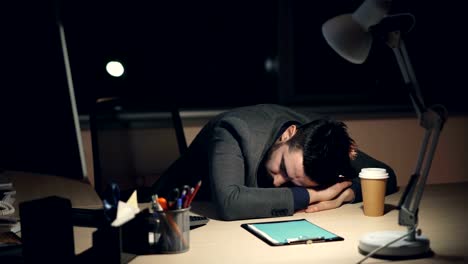 Tired-guy-in-suit-is-sleeping-on-work-desk-in-office-late-at-night-after-busy-working-day.-Turned-on-computer,-takeaway-coffee-and-lamp-are-visible-on-table.