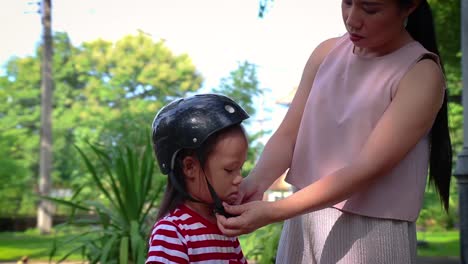 Madre-ayudando-a-su-hijo-(niño-de-pelo-largo)-a-ponerse-el-casco-de-la-bicicleta-al-aire-libre.