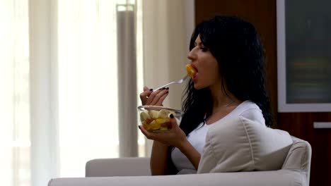 Joven-mujer-sentada-en-un-sofá-viendo-la-televisión-y-comer-ensalada-de-frutas-con-desayuno-saludable