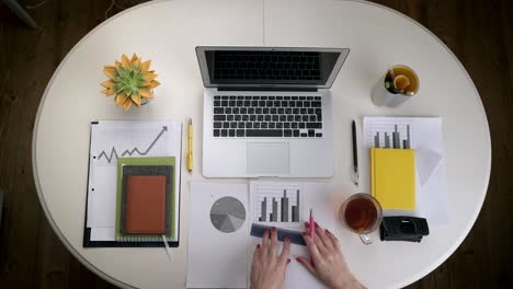 Woman-working-with-a-business-graph-and-a-laptop-that-stands-on-a-white-table-with-a-couple-tea.-Top-view.-Hands-close-up