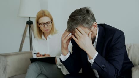 Upset-crying-businessman-patient-holding-his-head-by-hands-talking-to-his-psychoanalyst-in-her-office