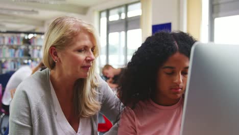 Teacher-With-Female-Student-Working-On-Computer-In-College-Library