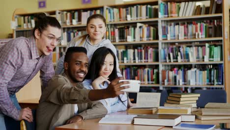 Grupo-de-estudiantes-internacionales-se-divierten-sonriendo-y-haciendo-fotos-selfie-en-cámara-del-smartphone-en-la-biblioteca-de-la-Universidad.-Amigos-alegres-tienen-descanso-mientras-preparan-para-examen