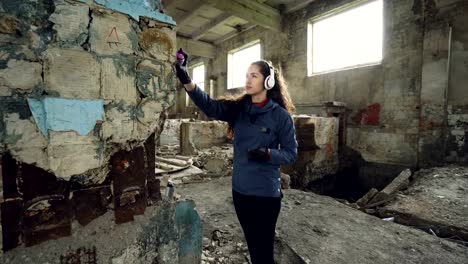 Pretty-young-woman-graffiti-artist-is-painting-with-spray-paint-inside-old-warehouse-and-listening-to-music-through-wireless-headphones.-Youth-subculture-concept.