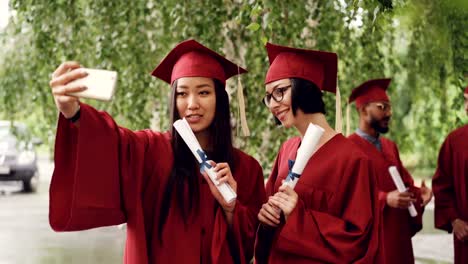 Chicas-guapas,-los-estudiantes-se-gradúan-son-selfie-toma-con-rollos-de-diploma-con-smartphone,-las-mujeres-plantean-y-sonriente,-sus-compañeros-están-celebrando-en-fondo.