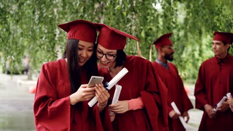 Female-graduates-are-using-smartphone-looking-at-screen-talking-and-laughing-standing-outdoors-holding-diplomas,-girls-are-wearing-formal-gowns-and-hats.