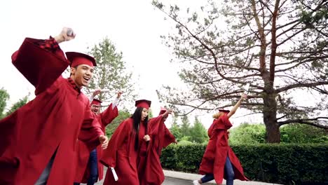 Slow-motion-of-merry-and-emotional-young-people-running-with-diplomas-wearing-mortar-boards-and-gowns-waving-scrolls-and-laughing.-Summertime-and-youth-concept.