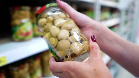 Manos-de-mujer-caucásica-de-Closeup-cerca-de-estantes-de-la-tienda-eligiendo-setas-escabeche-abarrotes-mercado