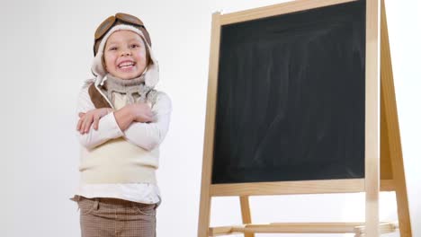 Una-pequeña-niña-vestida-como-un-aviador-o-piloto,-indica-con-su-mano-la-pizarra-detrás-de-ella-como-un-iniciar-vuelo-para-aprender-a-usar-el-avión-y-la-imaginación.