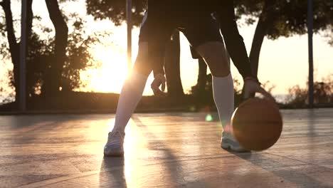 Close-up-footage-of-a-female-athlete-legs-in-white-golf-socks-and-sneakers.-Female-baasketball-player-bouncing-ball-from-hand-to-hand.,-practicing-cross-legs-exercise.-Sun-shines-on-the-background