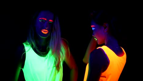 Women-with-UV-face-paint,-glowing-bracelets,-glowing-clothing-dancing-together-in-front-of-camera,-Half-body-shot.-Caucasian-and-asian-woman.-.