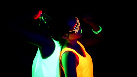 Women-with-UV-face-paint,-glowing-bracelets,-glowing-glasses,-clothing-dancing-back-against-each-other-in-front-of-camera,-Tilt-up-shot.-Caucasian-and-asian-woman.-Women.