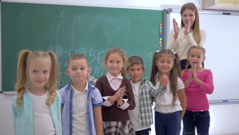 Grundschule,-Schulkinder-mit-Lehrer-in-die-Kamera-schauen-und-dann-auf-Hintergrund-von-Blackboard-applaudieren