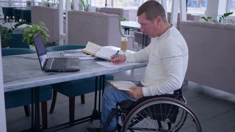 successful-diseased-student-man-on-wheel-chair-works-on-laptop-computer-during-distance-learning-for-online-education-and-makes-notes-in-notebook