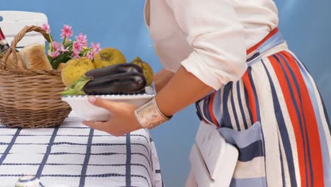 Mujer-joven-en-balcón-exterior-azul-con-bandeja-de-verduras.-Camisa-de-moda-blanco,-gran-sombrero-y-falda-colorida.-Tazas-de-café,-el-vino-y-el-pan-sobre-la-mesa.