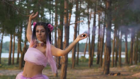 Beautiful-girl-with-bright-makeup-in-a-pink-dress-dancing-with-smoke-bombs-on-the-background-of-pine-trees.-The-dance-of-a-sensual-woman-with-a-flower-hairstyle.-Slow-motion.