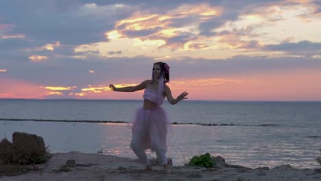 Linda-chica-con-maquillaje-brillante-en-un-vestido-rosa-bailando-en-la-orilla-del-río.-El-baile-de-una-mujer-sensual-con-un-peinado-maravilloso-con-flores.-Cámara-lenta.