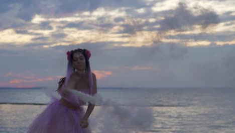 Tender-girl-with-sparkling-makeup-in-a-pink-dress-dancing-with-smoke-bombs-on-the-bank-of-the-river.-The-dance-of-a-sensual-woman-with-a-wonderful-hairstyle-with-flowers.-Slow-motion.