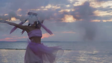 Hermosa-chica-con-maquillaje-brillante-en-un-vestido-rosado-bailando-con-bombas-de-humo-en-la-orilla-del-río.-El-baile-de-una-mujer-sensual-con-un-peinado-maravilloso-con-flores.-Cámara-lenta.