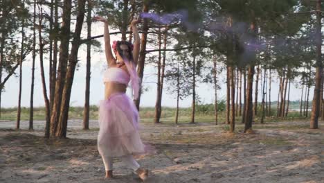 Charming-lady-with-bright-makeup-in-a-pink-dress-dancing-with-smoke-bombs-on-the-background-of-pine-trees.-The-dance-of-a-sensual-woman-with-a-flower-hairstyle.-Slow-motion.