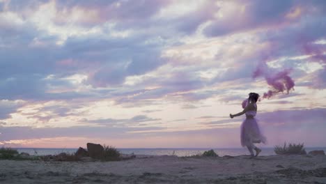 Linda-mujer-joven-con-maquillaje-brillante-en-un-vestido-rosado-bailando-con-bombas-de-humo-en-la-orilla-del-río.-El-baile-de-una-chica-sensual-con-un-peinado-maravilloso-con-flores.-Cámara-lenta.