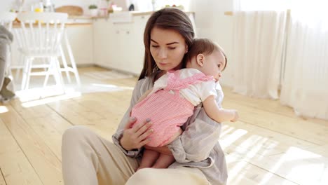 Liebevolle-junge-Mutter-tröstet-ihre-niedliche-Baby-Tochter-weinend-auf-ihrer-Schulter.-Frau-sitzt-zu-Hause-mit-ihrer-Tochter-in-den-Armen