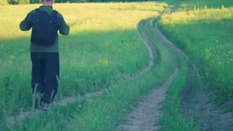 Un-mochilero-en-gorra-de-béisbol-azul,-chaqueta-de-vellón-verde,-pantalones-negros-y-zapatos-de-senderismo,-caminando-a-lo-largo-de-Downhill-Country-Dirt-Road-Winding-en-hierba-alta-al-atardecer.