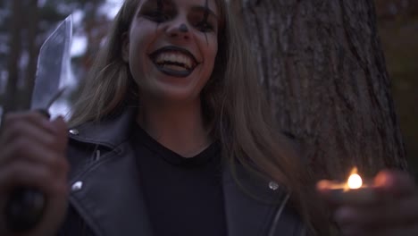 Crazy-young-woman-with-Halloween-clown-makeup-on-her-face-holding-big-knife-and-small-candle-in-hands