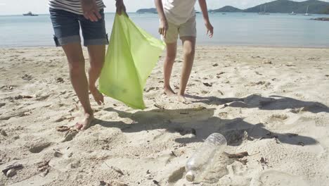 Mamá-y-su-hija-recogiendo-basura-en-la-playa-de-arena-en-una-bolsa-de-plástico-verde,-las-botellas-de-plástico-se-recogen-en-la-playa,-los-voluntarios-limpiando-la-playa.