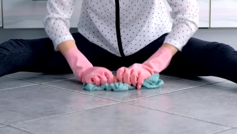 Woman-in-pink-rubber-gloves-washes-and-rubs-hard-the-stain-on-kitchen-floor-with-a-cloth.-Gray-tiles-on-the-floor.-Hands-close-up.