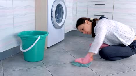 Mujer-cansada-con-guantes-de-goma-rosa-lava-y-frota-fuerte-la-mancha-en-el-suelo-de-la-cocina-con-un-paño.-Azulejos-grises-en-el-suelo.