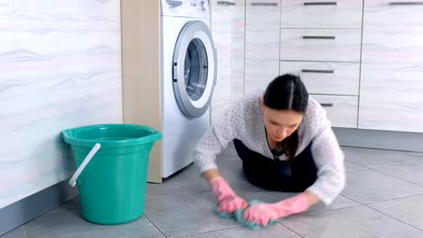 Tired-woman-in-pink-rubber-gloves-washes-kitchen-floor-with-a-cloth-and-looks-at-camera.-Gray-tiles-on-the-floor.