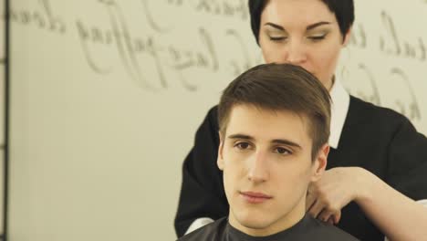 Frau-Friseur-trägt-Garderoben-Gewand-auf-jungen-Mann-vor-dem-Haarschnitt-im-Haarstudio.-Vorbereitung-für-Haarschnitt-in-Barber-Shop-Frong-Spiegel