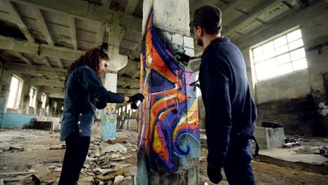 Low-angle-shot-of-graffiti-painters-decorating-abandoned-industrial-building-with-bright-images-using-aerosol-paint.-Dirty-old-walls-and-high-ceiling-are-visible.