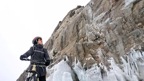 Man-is-walking-beside-bicycle-near-the-ice-grotto.-The-rock-with-ice-caves-and-icicles-is-very-beautiful.-The-cyclist-is-dressed-in-gray-down-jacket,-backpack-and-helmet.-The-traveler-is-ride-cycle.